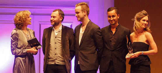 2009 Fünf Personen stehen auf einer Bühne, während eine Frau ein Mikrofon hält und spricht. Hintergrund in lila Beleuchtung.