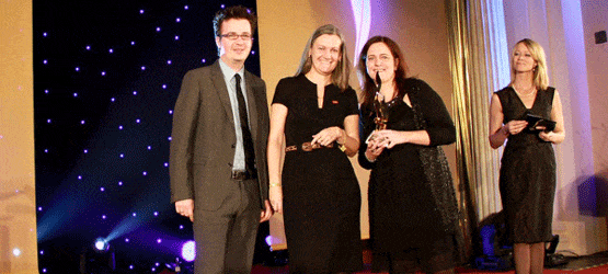 2010 Drei Personen stehen auf einer Bühne, lächeln und halten Auszeichnungen, während eine weitere Person im Hintergrund spr...