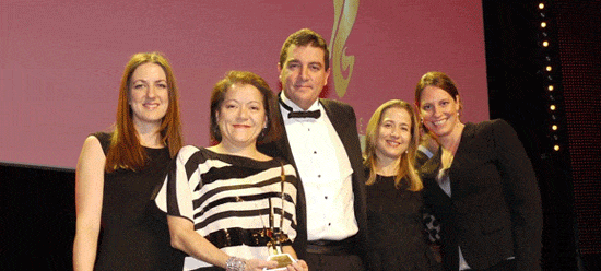 EEA_2012 Fünf Personen in formeller Kleidung stehen auf einer Bühne, eine hält einen Preis, Hintergrund mit rotem Banner.