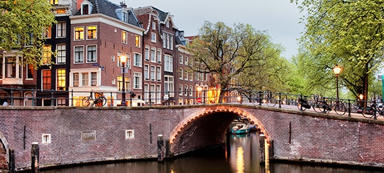 amsterdam-GettyImages-890686924 Brücke über einen Kanal in Amsterdam, umgeben von historischen Gebäuden und Bäumen, bei Dämmerung beleuchtet.