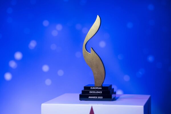 Goldene Auszeichnung auf einem Podest, beschriftet mit "European Excellence Awards 2023", vor einem blauen Hintergrund.