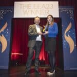 Zwei Personen auf einer Bühne halten einen Preis, umgeben von dekorativen Elementen und einem Banner mit "THE EEA23 WINNER".