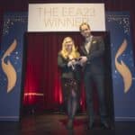 Zwei Personen auf einer Bühne halten einen Preis, umgeben von roten Vorhängen und einem Banner mit der Aufschrift "THE E...