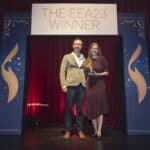 Zwei Personen stehen auf einer Bühne, halten eine Auszeichnung, vor einem Banner mit der Aufschrift "THE EEA23 WINNER".