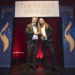 Zwei Männer in Anzügen stehen auf einer Bühne und halten eine Auszeichnung vor einem Banner mit der Aufschrift "THE EEA...