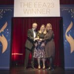 Drei Personen stehen auf einer Bühne, halten eine Auszeichnung, umgeben von rotem Vorhang und dekorativen Elementen.