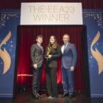 Drei Personen stehen auf einer Bühne vor einem Banner mit der Aufschrift "THE EEA23 WINNER" und halten eine Auszeichnung.