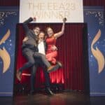 Drei Personen feiern auf einer Bühne, eine in rotem Kleid, zwei in Anzügen, mit einem Schild "THE EEA23 WINNER" im Hinte...