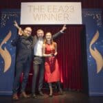 Drei Personen posieren auf einer Bühne vor einem Banner mit der Aufschrift "THE EEA23 WINNER" und halten Auszeichnungen.