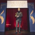 Mann in eleganter Kleidung hält einen Preis vor einem roten Vorhang und einem Banner mit der Aufschrift "THE EEA23 WINNER".