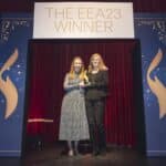Zwei Frauen stehen auf einer Bühne, halten einen Preis, vor einem Banner mit der Aufschrift "THE EEA23 WINNER".