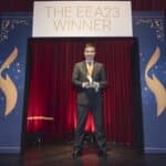 Mann in Anzug hält Trophäe vor einem Banner mit der Aufschrift "THE EEA23 WINNER" auf rotem Vorhang.