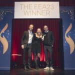 Drei Personen stehen vor einem Bühnenhintergrund mit der Aufschrift "THE EEA23 WINNER" und feiern einen Preis.