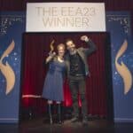 Zwei Personen stehen vor einem Siegerbanner, eine hält einen Preis, beide lächeln und posieren freudig.