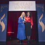 Zwei Frauen stehen auf einer Bühne vor einem Banner mit der Aufschrift "THE EEA23 WINNER", umgeben von rotem Vorhang.