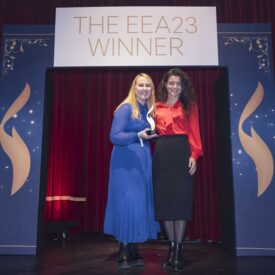 Zwei Frauen stehen auf einer Bühne vor einem Banner mit der Aufschrift "THE EEA23 WINNER", umgeben von rotem Vorhang.