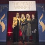 Vier Frauen stehen auf einer Bühne vor einem Banner mit der Aufschrift "THE EEA23 WINNER" in einem festlichen Raum.