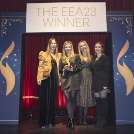 Vier Frauen stehen auf einer Bühne vor einem Banner mit der Aufschrift "THE EEA23 WINNER" in einem festlichen Raum.