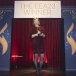 Frau in rotem, glitzerndem Kleid hält einen Preis auf einer Bühne mit rotem Vorhang und "THE EEA23 WINNER"-Schild.