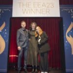 Drei Personen stehen auf einer Bühne vor einem roten Vorhang, mit einem Schild "THE EEA23 WINNER" im Hintergrund.