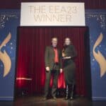 Zwei Personen stehen auf einer Bühne vor einem Banner mit der Aufschrift "THE EEA23 WINNER". Rote Vorhänge im Hintergrund.