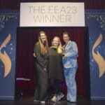 Drei Frauen stehen lächelnd vor einem Banner mit der Aufschrift "THE EEA23 WINNER" auf einer Bühne.