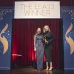 Zwei Frauen stehen auf einer Bühne vor einem Banner mit der Aufschrift "THE EEA23 WINNER" und halten eine Auszeichnung.