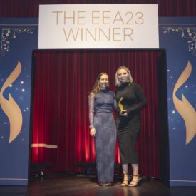 Zwei Frauen stehen auf einer Bühne vor einem Banner mit der Aufschrift "THE EEA23 WINNER" und halten eine Auszeichnung.