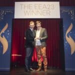 Zwei Männer stehen auf einer Bühne, halten eine Auszeichnung, umgeben von rotem Vorhang und einem Banner mit "THE EEA23 ...