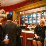 Zwei Frauen in eleganter Abendkleidung sitzen an einer Bar, umgeben von festlicher Dekoration und Gästen.