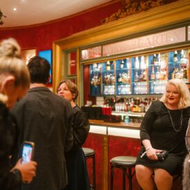 Zwei Frauen in eleganter Abendkleidung sitzen an einer Bar, umgeben von festlicher Dekoration und Gästen.