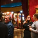 Menschen in eleganter Umgebung, einige halten Gläser, mit rotem Hintergrund und Kunstwerken an den Wänden.