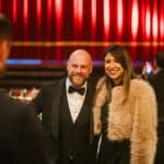 Paar in eleganter Abendgarderobe lächelt vor rotem Vorhang, festliche Atmosphäre.