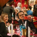 Zwei Personen machen ein Selfie in einem festlich dekorierten Restaurant mit vielen Gästen im Hintergrund.