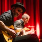 Musiker spielt Gitarre auf der Bühne, rote Vorhänge im Hintergrund, ein weiterer Musiker im Hintergrund sichtbar.