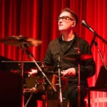 Mann mit Brille spielt Schlagzeug vor rotem Vorhang auf einer Bühne.