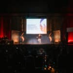 Bühne mit Redner bei den European Excellence Awards 2023, umgeben von Publikum und festlicher Beleuchtung.