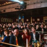 Publikum in einem Theater, aufmerksam auf die Bühne, mit Tischen und Kerzenlicht im Vordergrund.