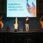 Zwei Moderatoren stehen auf einer Bühne vor einer Leinwand mit dem Text "European Excellence Awards 2023".