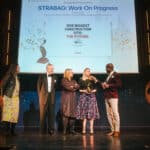 Vier Personen stehen auf einer Bühne, während ein Mann eine Auszeichnung hält. Im Hintergrund ist eine Präsentation si...