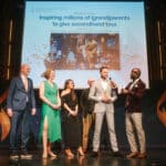 Eine Preisverleihung mit mehreren Personen auf der Bühne, einem Präsentator und einer Projektionsleinwand im Hintergrund.