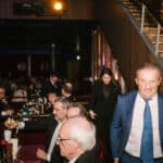 Mann in Anzug geht lächelnd durch einen eleganten Veranstaltungsraum mit Tischen und Gästen.