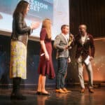 Vier Personen auf einer Bühne bei einer Preisverleihung, eine Person hält einen Preis, festliche Atmosphäre.