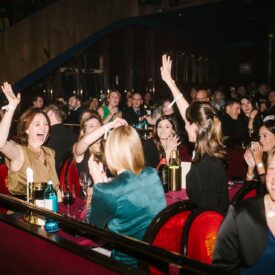 Menschen in einem Veranstaltungsraum applaudieren und feiern, mit Lichtern und Tischen im Hintergrund.