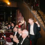 Menschen in eleganter Kleidung applaudieren an einem Tisch, während ein Mann jubelnd aufsteht. Treppe im Hintergrund.