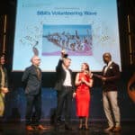 Zwei Personen halten einen Preis, während drei weitere Personen auf einer Bühne stehen, im Hintergrund eine Präsentation.