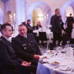 Zwei Männer in Uniform sitzen an einem festlich gedeckten Tisch, umgeben von Gästen in einem eleganten Veranstaltungsraum.