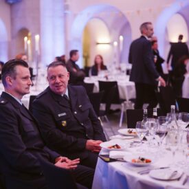 Zwei Männer in Uniform sitzen an einem festlich gedeckten Tisch, umgeben von Gästen in einem eleganten Veranstaltungsraum.