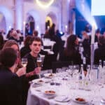 Junge Menschen heben Gläser in einem festlich gedeckten Saal mit Tischen und Kerzenlicht bei einer Veranstaltung.