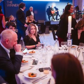 Elegantes Event mit Gästen an Tischen, einem Musiker im Hintergrund und festlicher Atmosphäre.